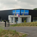 Vue extérieure du magasin BE SPA UNIVERS à Barentin, partageant l'espace avec Desjoyaux Piscines. Le bâtiment présente une façade moderne noire et blanche avec de grandes enseignes lumineuses pour chaque marque. Un parking spacieux est disponible devant le magasin, offrant un accès facile pour les clients.