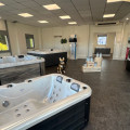 Showroom de Cosy Piscine et Paysage Savigneux présentant une sélection de jacuzzis et spas de nage. L'espace est lumineux avec de grandes fenêtres, un sol en vinyle gris et des murs décorés de manière contemporaine. Des statues décoratives et des produits d'entretien pour piscine sont également exposés, offrant une ambiance accueillante et professionnelle pour les clients.