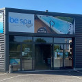 Vue extérieure du magasin BE SPA Ormes - Eau et bulles, affichant une façade moderne avec des enseignes grandes et visibles pour les spas, les piscines et les services. Le design élégant en métal ondulé est complété par des affichages publicitaires lumineux et une fontaine décorative à l'entrée, offrant un accueil attrayant aux visiteurs.