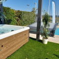 Showroom extérieur d'Evasion Piscines à Saint-Maximin-la-Sainte-Baume, mettant en scène un jacuzzi moderne sur un socle en bois avec un gazon artificiel. L'espace est élégamment aménagé avec des plantes décoratives, à côté d'une piscine visible en arrière-plan, sous un ciel clair, reflétant l'expertise de l'entreprise dans la création d'espaces de détente extérieurs.