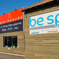Façade moderne du magasin JV Pose à Saint Andiol, combinant des services de menuiserie et la vente de spas BE SPA. Le bâtiment présente une grande enseigne rouge 'SUR-MESURE JV Pose Menuiserie Bois - PVC - Alu' à côté d'un panneau 'be spa France Créateur de bien-être'. Des plantes en pots accueillent les visiteurs à l'entrée, reflétant un engagement envers le bien-être et la qualité artisanale.