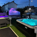 xposition extérieure nocturne chez Piscine & Spa Service 52 à Foulain, présentant des spas BE SPA éclairés. Deux jacuzzis sont mis en valeur avec des lumières LED multicolores sous un ciel crépusculaire. Un panneau informatif est placé à côté de chaque spa, offrant des détails sur les modèles et leurs caractéristiques. L'installation est sur un gazon synthétique, avec un éclairage d'ambiance qui souligne l'élégance des spas