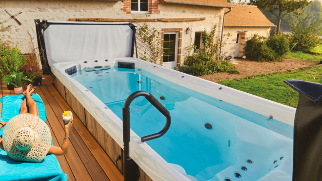 Image d'une femme allongée détendue sur une chaise longue sur une terrasse, à côté d'un spa de nage. La scène dépeint un moment de relaxation en plein air, avec la femme profitant du soleil et de la tranquillité environnante, évoquant un style de vie sain et relaxant.