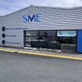 Façade du magasin SME Eau & Bien-être à Vaux Sur Mer, montrant un bâtiment industriel avec un grand logo SME en bleu sur le toit. La vitrine du magasin affiche clairement le logo de BE SPA, indiquant qu'il est distributeur de spas de cette marque. L'extérieur du magasin présente un design moderne et accueillant, avec des affiches de promotion des bienfaits de l'eau et du bien-être.
